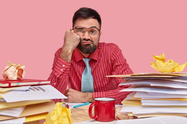 Displeased, tired male worker keeps fist on cheek, has sleepy look, dressed in shirt and tie, has too much admin on desk